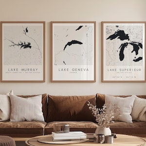 May include: Three framed prints of lake maps hang above a brown sofa with white and brown pillows. The prints feature the names Lake Murray, Lake Geneva, and Lake Superior. A round wooden coffee table and ottoman are in front of the sofa.