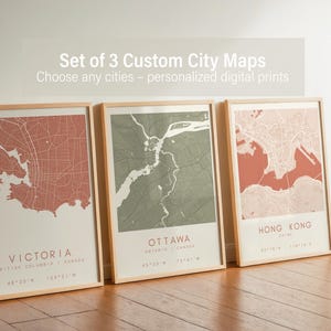 May include: Three framed city maps with a light wood frame. The maps are in shades of red, green, and coral. The text at the top says "Set of 3 Custom City Maps". The cities are Victoria, Ottawa, and Hong Kong.
