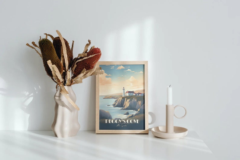 K&ouml;nnte beinhalten: Ein gerahmter Druck eines Leuchtturms auf einer Klippe mit Blick auf das Meer. Der Druck tr&auml;gt den Titel "Peggy's Cove, Nova Scotia". Der Druck wird auf einer wei&szlig;en Oberfl&auml;che mit einer Vase mit getrockneten Blumen und einem Kerzenhalter angezeigt.