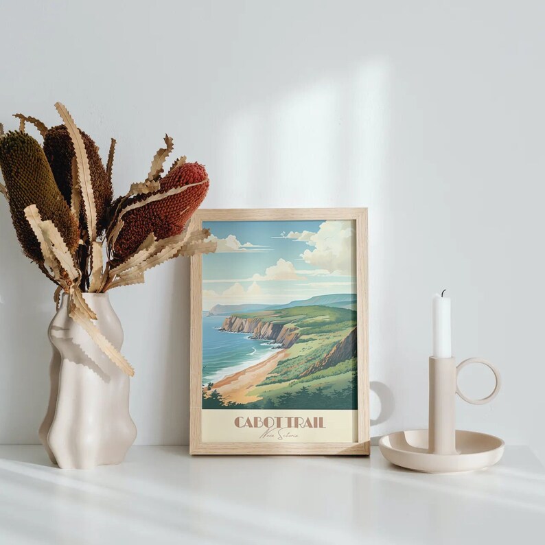 May include: A framed print of a coastal landscape with the text "Cabot Trail Nova Scotia" in a light brown frame. The print is on a white surface with a vase of dried flowers to the left and a candle holder to the right.