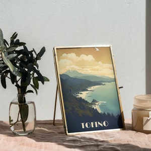 K&ouml;nnte beinhalten: Ein goldfarbener Bilderrahmen mit einem Vintage-Reiseposter eines K&uuml;stenlandschafts. Das Poster zeigt ein blaues Meer, gr&uuml;ne Berge und das Wort "TOFINO" in wei&szlig;er Schrift. Der Rahmen steht auf einer hellrosa Oberfl&auml;che mit einer Vase mit gr&uuml;nen Bl&auml;ttern und einer beigefarbenen Kerze.