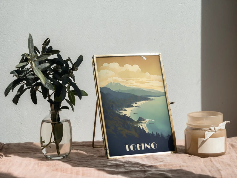 K&ouml;nnte beinhalten: Ein goldfarbener Bilderrahmen mit einem Vintage-Reiseposter eines K&uuml;stenlandschafts. Das Poster zeigt ein blaues Meer, gr&uuml;ne Berge und das Wort "TOFINO" in wei&szlig;er Schrift. Der Rahmen steht auf einer hellrosa Oberfl&auml;che mit einer Vase mit gr&uuml;nen Bl&auml;ttern und einer beigefarbenen Kerze.