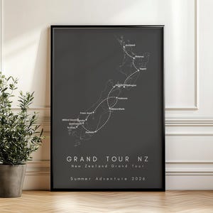 May include: A framed print featuring a map of New Zealand in black and white, with a dotted route. The text reads "GRAND TOUR NZ" and "Summer Adventure 2026". A potted plant is in the foreground, adding a touch of greenery.