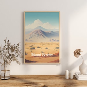 May include: A framed poster featuring a vintage travel illustration of Mount Bromo in Indonesia. The poster depicts a blue sky with white clouds, a brown mountain with a green peak, and a desert landscape with brown sand and green bushes. The text "Mount Bromo" is printed in white on the poster.