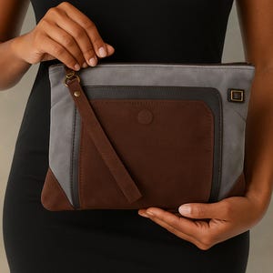 May include: A multi-tone clutch with a brown, gray, and dark gray color scheme. The clutch features a rectangular shape with a brown pocket and a wrist strap. The design includes a button closure and a metal buckle detail.