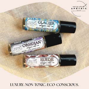 以下が含まれることがあります： 黒いキャップが付いた3つのロールオン香水ボトルが表示されています。「Glacier」、「Obsidian」、「Suede」と書かれたラベルには香りの説明があります。ボトルはベージュ色の布の表面にあります。「Luxury. Non-Toxic. Eco-Conscious.」というテキストが下にあります。