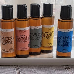 Könnte beinhalten: Fünf kleine Glasflaschen mit braunen Kappen und Etiketten. Die Etiketten sind weiß mit schwarzem Text und zeigen verschiedene Düfte: Dirty Chai, Kiss, Midnight, Moon Flower und Volcanic. Die Flaschen sind in einer Reihe auf einer weißen Oberfläche angeordnet.