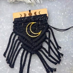May include: Black macrame wall hanging with a gold crescent moon accent. The top is a wooden piece with moon phase cutouts. The macrame has multiple knotted strands. The background is a light gray surface.
