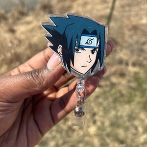 Puede incluir: Un portagafete de plástico transparente con una cara de personaje de anime negro y azul. El personaje tiene el pelo negro, los ojos azules y una diadema con un símbolo de espiral.