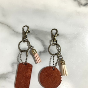 Può includere: Due portachiavi con ciondoli in pelle marrone, uno rettangolare e uno rotondo. Ogni ciondolo ha un anello in metallo dorato e un piccolo pon pon. I pon pon sono di colori diversi, uno è rosa e l'altro è bianco.