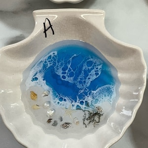 May include: A white, seashell-shaped dish featuring a blue and white resin ocean wave design. The dish is adorned with tiny seashells, a starfish, and sand-like details. The letter "A" is written in black on the dish.