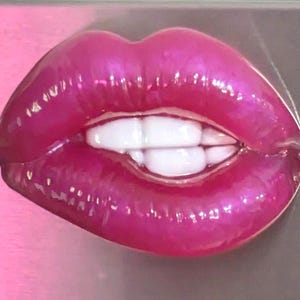 May include: A glossy, vibrant magenta-coloured brooch shaped like a pair of lips. The lips are depicted in a biting pose, revealing a set of smooth, white teeth tucked behind the lower lip. The surface has a reflective, high-shine finish.