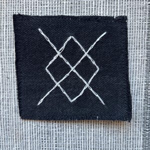 May include: A black fabric patch with a white embroidered diamond shape with intersecting lines.