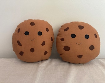 Puede incluir: Dos cojines redondos marrones con forma de galleta, con detalles bordados de chispas de chocolate y caras sonrientes. Los cojines están hechos de una tela suave y texturizada, que se asemeja a galletas con chispas de chocolate.