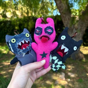 May include: Three handmade monster plush toys. The toys are held in a hand. The plush toys are in shades of pink, black, and denim blue. The pink monster has a green star and a tongue sticking out. The other two have open mouths with white teeth.
