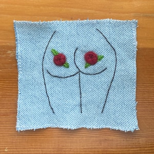 May include: A blue denim patch with a simple embroidered outline of a pair of buttocks with red roses on each cheek.