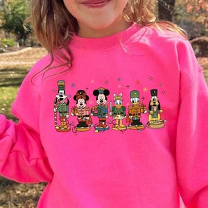 May include: A bright pink sweatshirt featuring a graphic of cartoon nutcrackers. The nutcrackers are dressed in various outfits and stand in a row. The sweatshirt is worn by a young person.