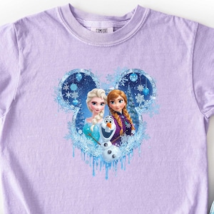 May include: A lavender t-shirt featuring a Disney-themed graphic of Elsa and Anna from Frozen, within a Mickey Mouse-shaped design. The shirt is paired with light blue denim shorts and black and white checkered slip-on shoes.