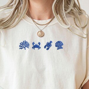 May include: Cream-colored t-shirt with blue embroidered sea-themed designs, including a coral, crab, and seashell. The shirt is part of the "Comfort Colors" collection and is a personalized gift.