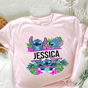 May include: Pink t-shirt with a graphic of three blue Stitch characters from Lilo & Stitch, surrounded by tropical leaves and pink flowers. The text "JESSICA" is printed in black above the characters.