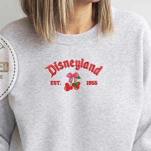 May include: Gray sweatshirt with a white embroidered design that says "Disneyland Est. 1955" with a red bow and cherries.
