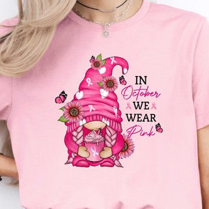 May include: Pink t-shirt with a pink gnome holding a pink cup with a pink ribbon. The gnome is wearing a pink hat with a pink ribbon and has pink hair. The shirt has the text "In October We Wear Pink" on it.