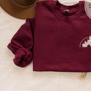 Puede incluir: Sudadera color burdeos con una imagen bordada en blanco del castillo de Disney en la parte delantera. La sudadera est&aacute; doblada y colocada sobre una superficie de color marr&oacute;n claro.