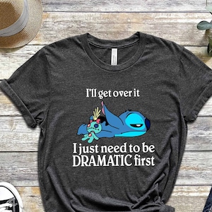 May include: A dark gray t-shirt with a white graphic of Stitch from Lilo & Stitch. The graphic shows Stitch lying down with a sad expression and the text "I'll get over it I just need to be DRAMATIC first".