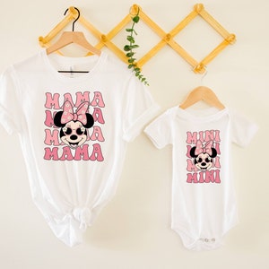 Pu&ograve; includere: Maglietta bianca con una grafica rosa di Minnie Mouse e il testo "MAMA" ripetuto quattro volte. Un body bianco con una grafica rosa di Minnie Mouse e il testo "MINI" ripetuto quattro volte.