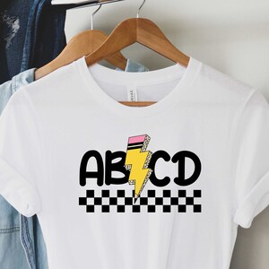 May include: White t-shirt with a black and yellow lightning bolt graphic and the text "ABCD" in black. The lightning bolt has a pink eraser on top. A black and white checkered pattern is below the text.