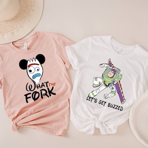 May include: Two t-shirts with a pink and white color scheme. The pink t-shirt features a cartoon image of a fork with Mickey Mouse ears and the text "What the Fork". The white t-shirt features a cartoon image of Buzz Lightyear from Toy Story and the text "Let's get buzzed".