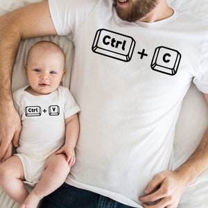 May include: A white t-shirt with the text "Ctrl + C" printed on it. A white onesie with the text "Ctrl + V" printed on it. A father and baby are wearing the shirts.