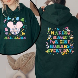 Puede incluir: Sudadera con capucha verde oscuro con un dise&ntilde;o colorido que presenta un castillo, orejas de Mickey Mouse y el texto "MRS. BROWN" en la parte delantera. La parte posterior de la sudadera dice "MAKING MAGIC FOR TINY HUMANS EVERY DAY" con personajes de dibujos animados.