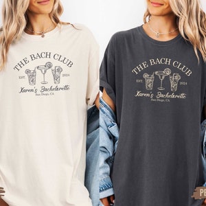 Puede incluir: Dos camisetas con un dise&ntilde;o de despedida de soltera. La camiseta de color marfil y la camiseta gris oscuro presentan el texto "THE BACH CLUB EST. 2024 Karen's Bachelorette San Diego, CA" y un gr&aacute;fico de tres vasos de c&oacute;ctel.