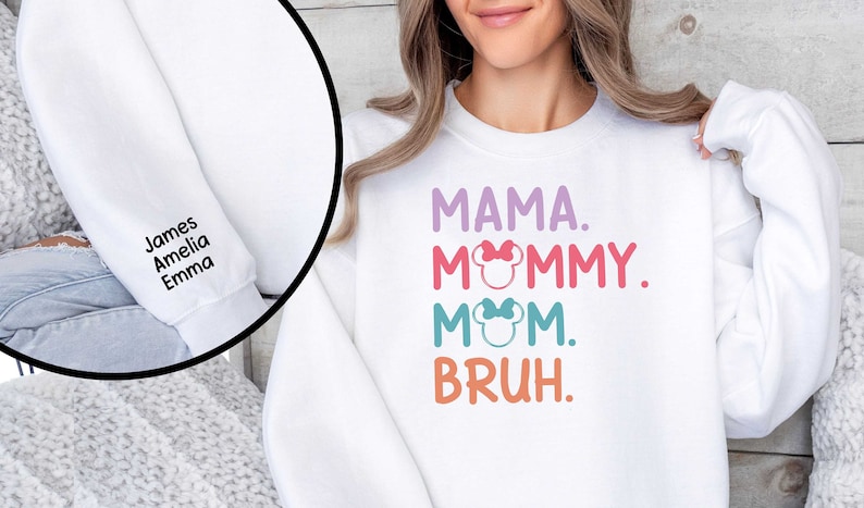 K&ouml;nnte beinhalten: Wei&szlig;er Pullover mit dem Text "MAMA. MOMMY. MOM. BRUH." in bunten Buchstaben mit einem Minnie Mouse-Kopf-Motiv. Der &Auml;rmel hat die Namen "James, Amelia, Emma" in Schwarz gestickt.