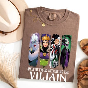 Pu&ograve; includere: Una maglietta marrone con quattro personaggi di cattivi dei cartoni animati e la scritta "Sweetie I'm OK with Being the Villain". L'immagine include un cappello, sandali e una collana d'oro.