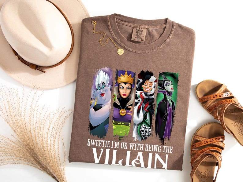 Pu&ograve; includere: Una maglietta marrone con quattro personaggi di cattivi dei cartoni animati e la scritta "Sweetie I'm OK with Being the Villain". L'immagine include un cappello, sandali e una collana d'oro.