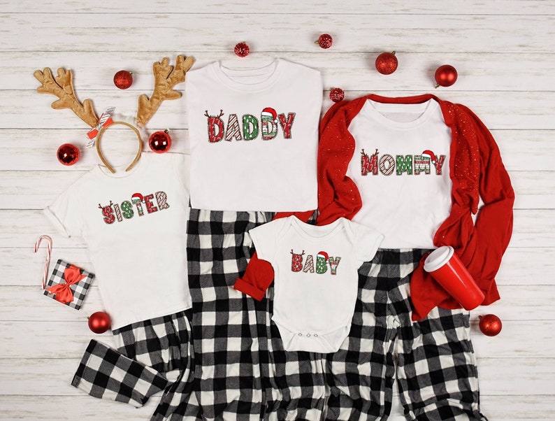 May include: A family of four wearing matching Christmas pajamas. The pajamas are white with red and green plaid trim. The shirts have a Christmas-themed design with the words "Daddy", "Mommy", "Sister", and "Baby".