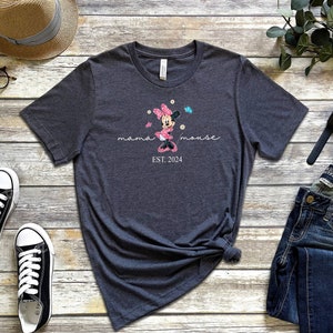 Op de afbeelding: Een donkergrijs T-shirt met een roze en witte Minnie Mouse-afbeelding, de tekst "mama mouse EST. 2024" en een paar vlinders.