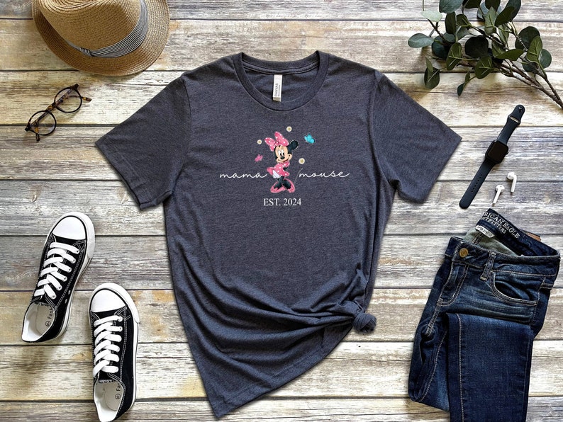 Op de afbeelding: Een donkergrijs T-shirt met een roze en witte Minnie Mouse-afbeelding, de tekst "mama mouse EST. 2024" en een paar vlinders.