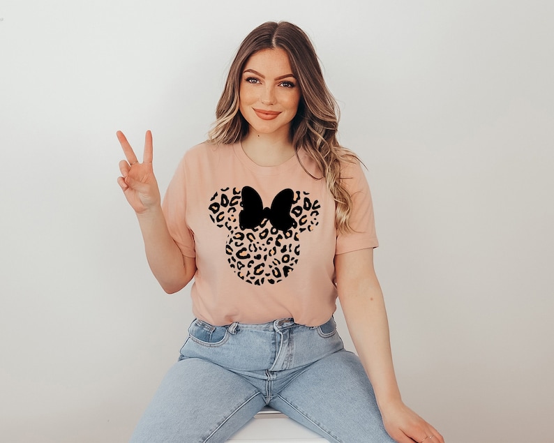 May include: A light pink t-shirt with a black and brown leopard print Mickey Mouse head design. The design features a black bow.