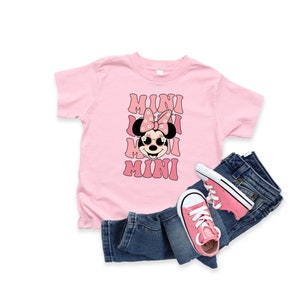 Pu&ograve; includere: Maglietta rosa con un'immagine di Minnie Mouse che indossa occhiali da sole e un fiocco con fiori. Il testo "MINI" &egrave; ripetuto quattro volte in un font retr&ograve;.