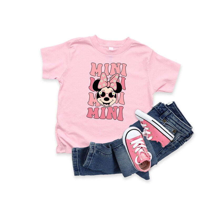 Pu&ograve; includere: Maglietta rosa con un'immagine di Minnie Mouse che indossa occhiali da sole e un fiocco con fiori. Il testo "MINI" &egrave; ripetuto quattro volte in un font retr&ograve;.