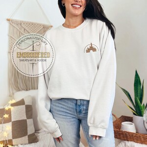 Puede incluir: Sudadera blanca con una imagen bordada en dorado de un castillo en el pecho izquierdo. La sudadera tiene cuello redondo y mangas largas. El texto "Regalo personalizado" y "Sudadera y sudadera con capucha bordadas" est&aacute; en un c&iacute;rculo blanco con un contorno dorado.