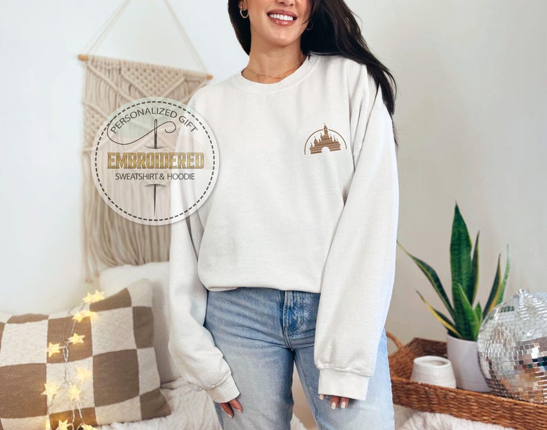 Puede incluir: Sudadera blanca con una imagen bordada en dorado de un castillo en el pecho izquierdo. La sudadera tiene cuello redondo y mangas largas. El texto "Regalo personalizado" y "Sudadera y sudadera con capucha bordadas" est&aacute; en un c&iacute;rculo blanco con un contorno dorado.
