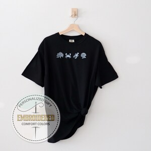 May include: Black t-shirt with embroidered light blue sea-themed designs: seaweed, crab, lobster, and seashell. The shirt is hanging on a wooden hanger against a white background. The text "EMBROIDERED COMFORT COLORS" is on a circular label.