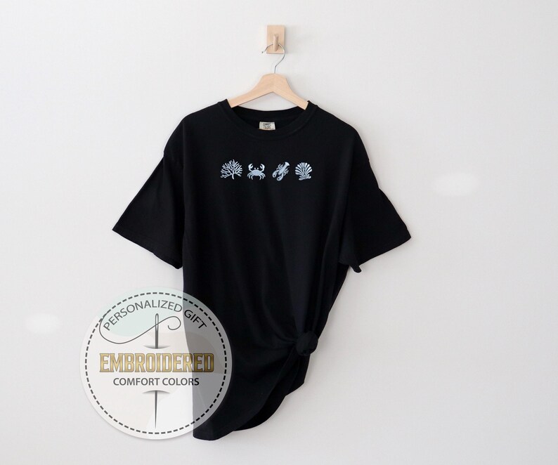 May include: Black t-shirt with embroidered light blue sea-themed designs: seaweed, crab, lobster, and seashell. The shirt is hanging on a wooden hanger against a white background. The text "EMBROIDERED COMFORT COLORS" is on a circular label.