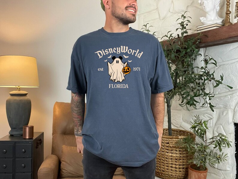 May include: A dark blue t-shirt featuring a white graphic of a ghost holding a jack-o-lantern. The text "DisneyWorld est 1971 FLORIDA" is printed on the shirt.