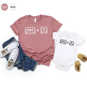 May include: A heather mauve t-shirt with the text "Ctrl + C" in white, and a white bodysuit with the text "Ctrl + V" in black. The t-shirt is paired with blue jeans and white sneakers.