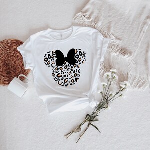 May include: White t-shirt with a black and orange leopard print Minnie Mouse head design. The bow is black.